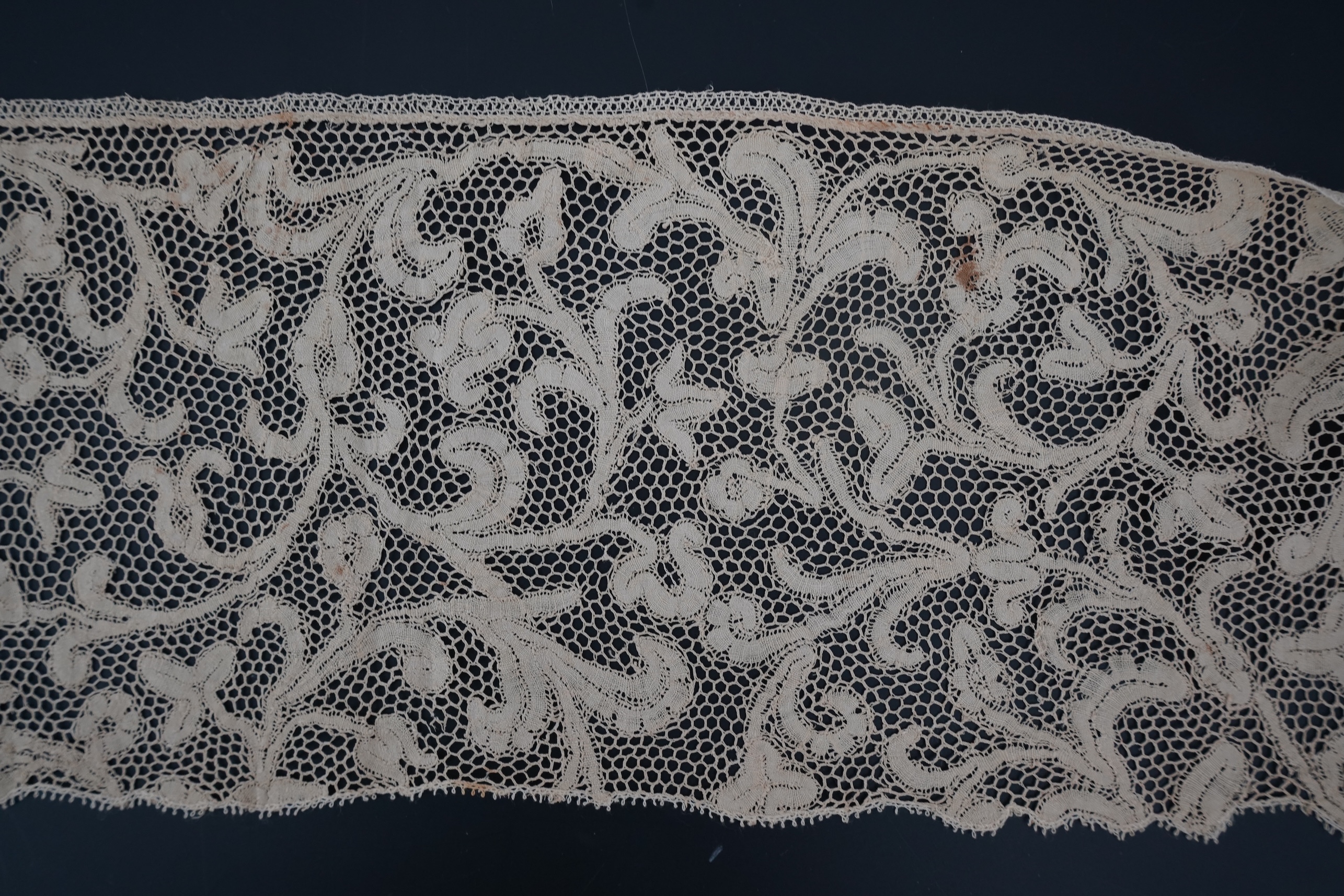 A length of 1740-1760 Italian, Milanese bobbin lace flounce, made from flat braiding or tapes, placed into scrolling patterns, attached to brides, made into a honeycomb mesh. 252cm long x 16cm deep. Condition - one small